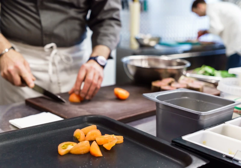 Chef slicing tomatoes on a cutting board in a commercial kitchen with food prep containers nearby.