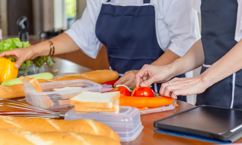 Two people preparing sandwiches with bread, tomatoes, carrots, and fresh vegetables on a kitchen table.