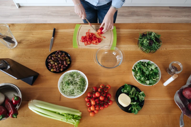 Overhead view of a person slicing red peppers with fresh salad ingredients arranged on a wooden table.